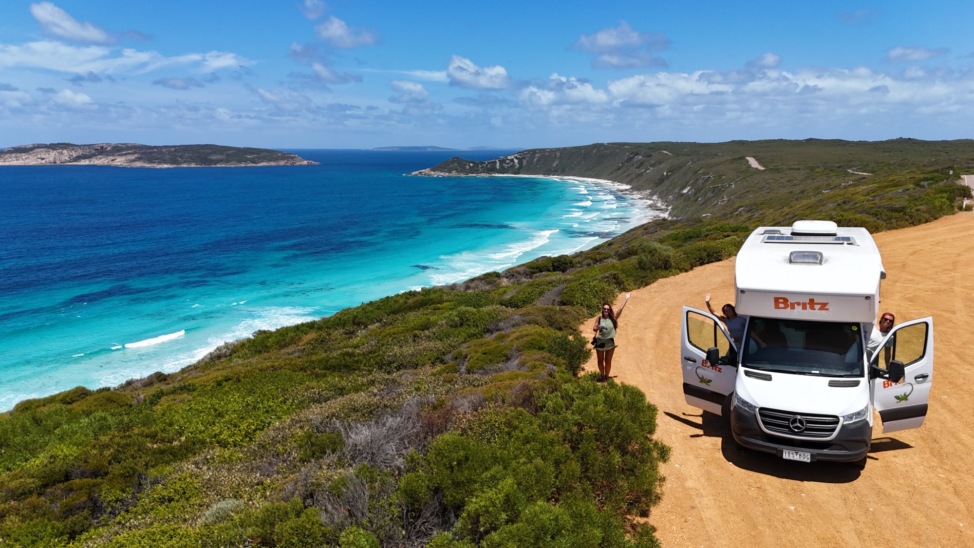 Three WanderBuddies members waving beside their hired Britz campervan above a turquoise bay near Esperance, Western Australia